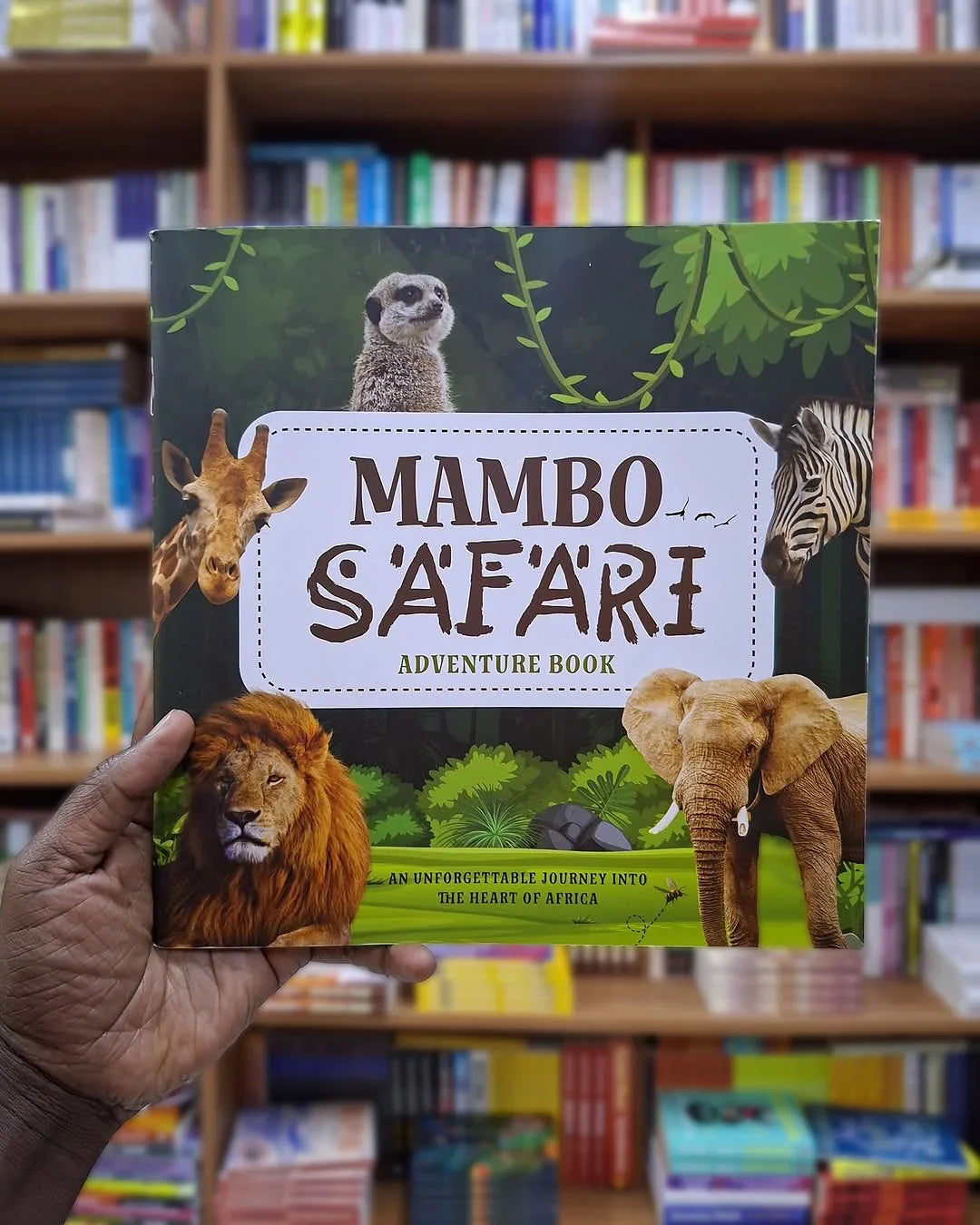 Mambo Safari Adventure Book cover featuring lion, elephant, giraffe, zebra, and meerkat with African jungle background, held in a bookstore.